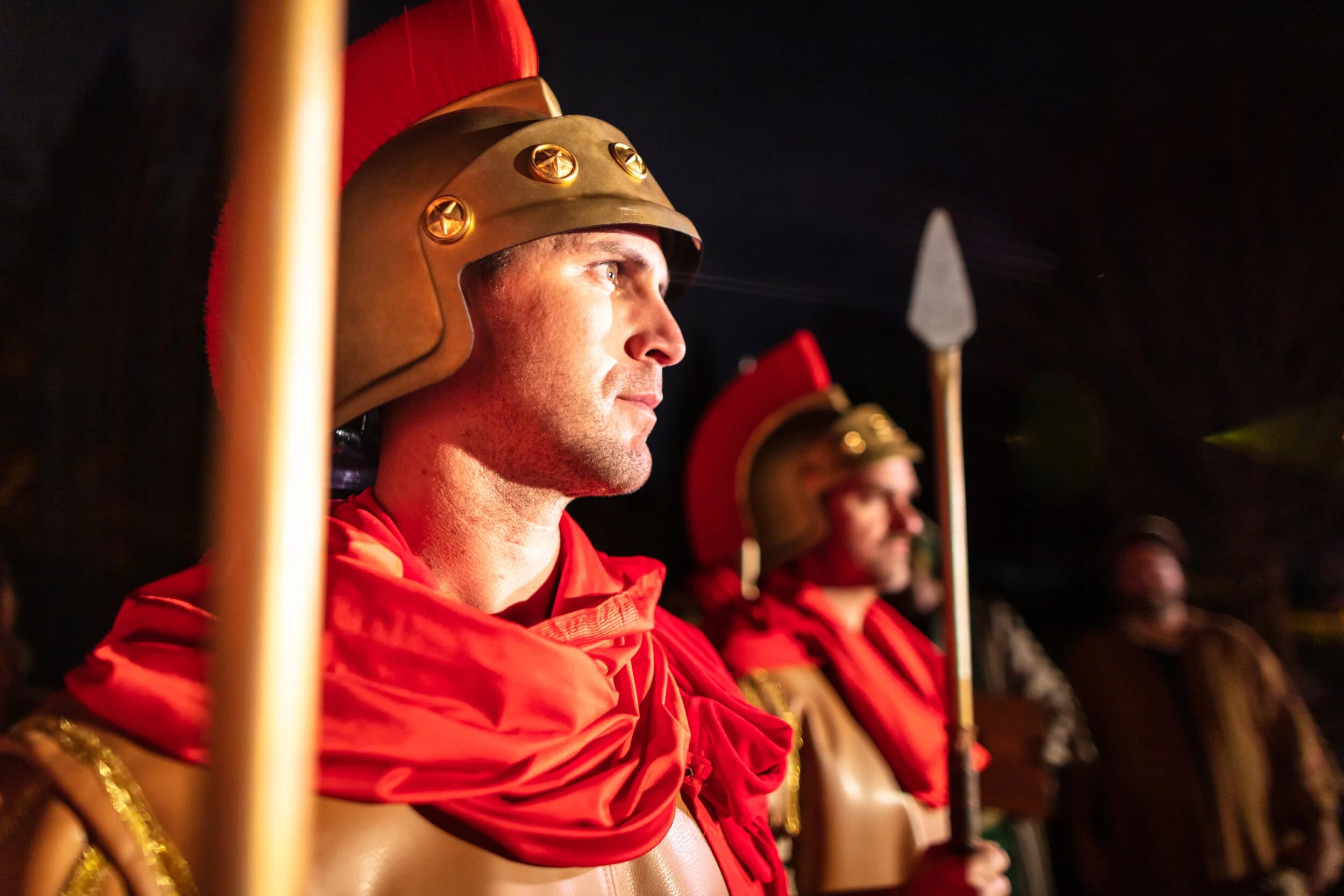A Roman soldier in profile wears a bronze crested helmet with a red plume and cape, holding a spear beside another soldier.