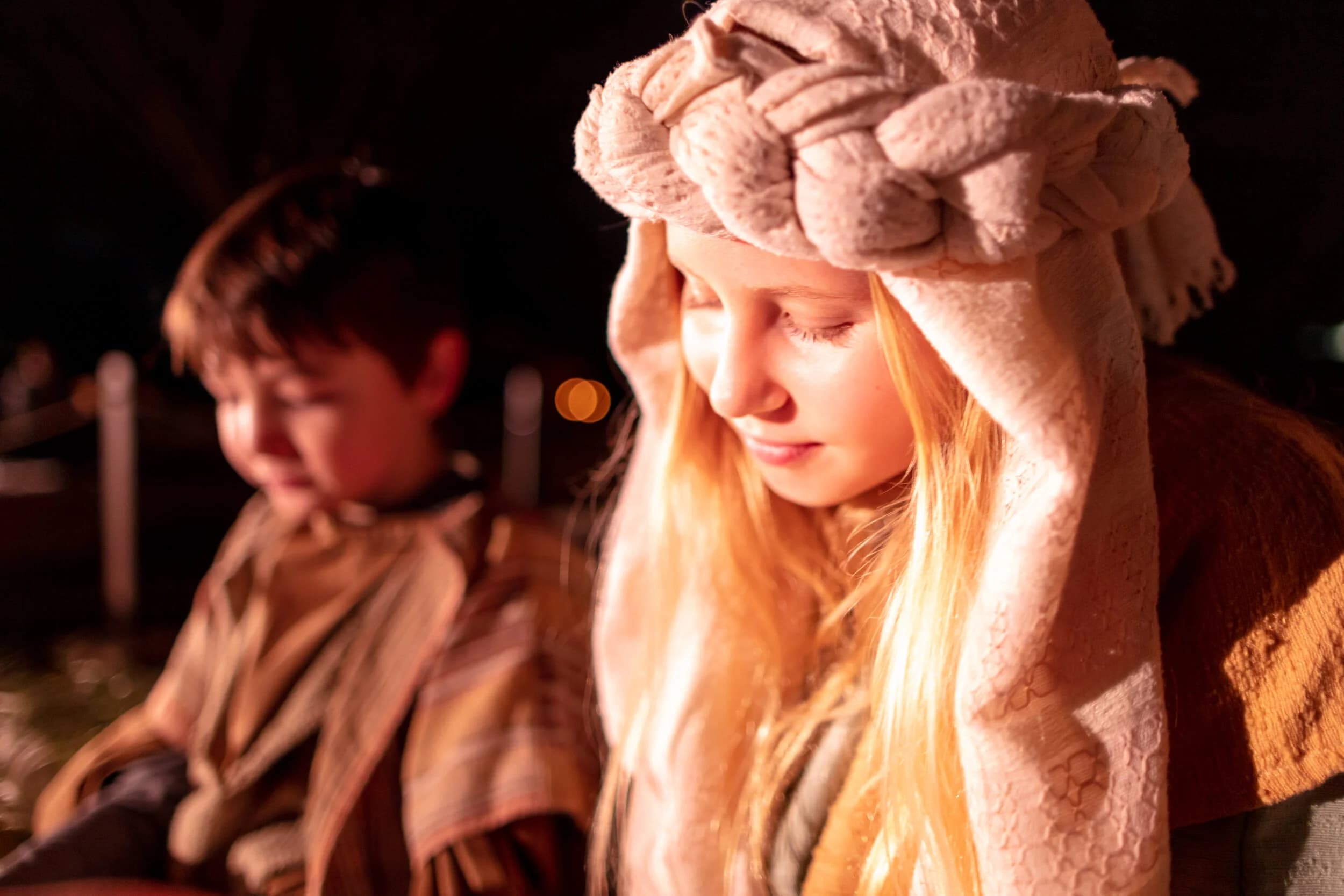 A girl adjusts her cream headscarf in firelight while a boy in costume stands blurred behind her at night.