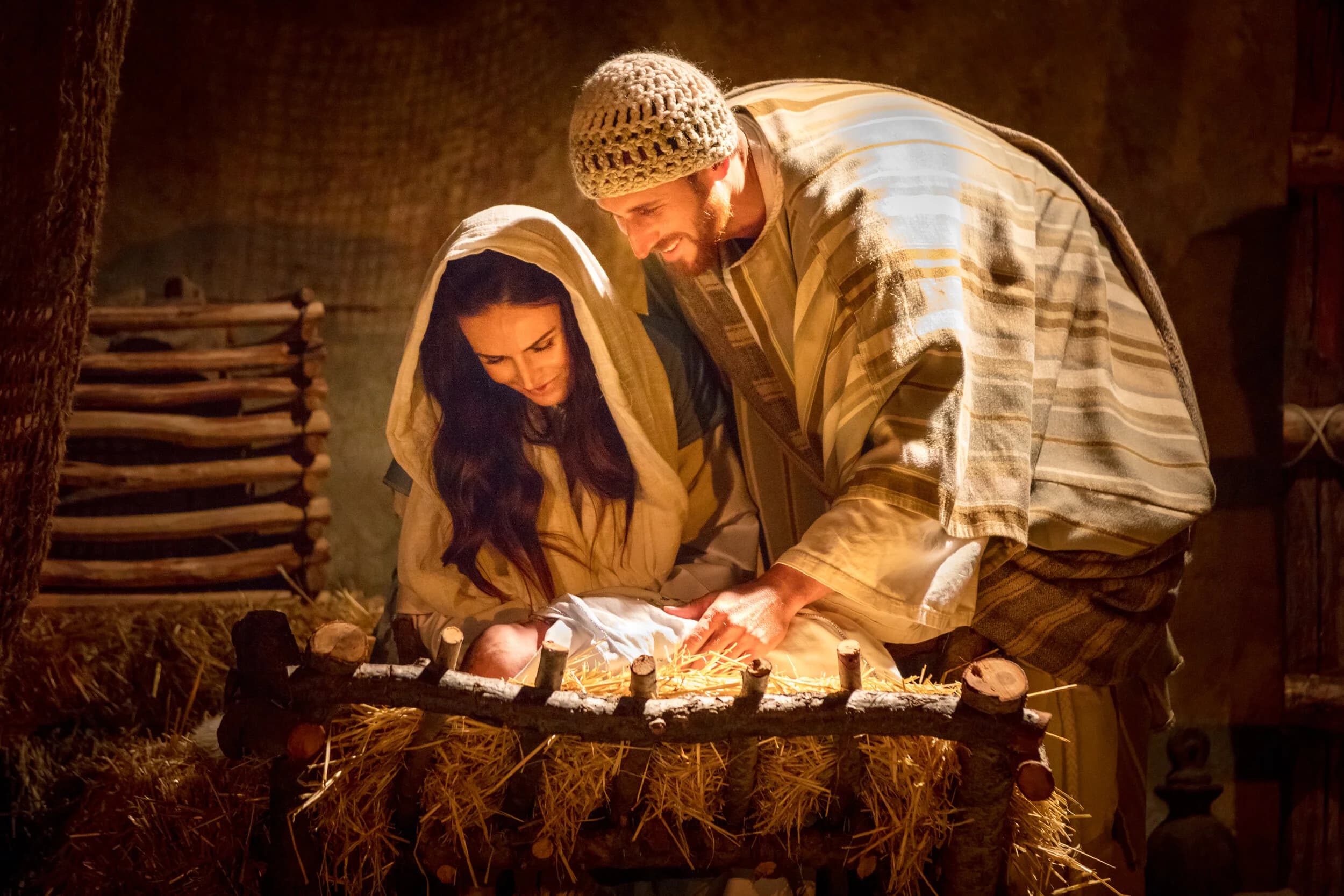 Joseph in a striped robe and knit cap leans over Mary as she tends the baby in a hay lined manger inside a wooden stable.