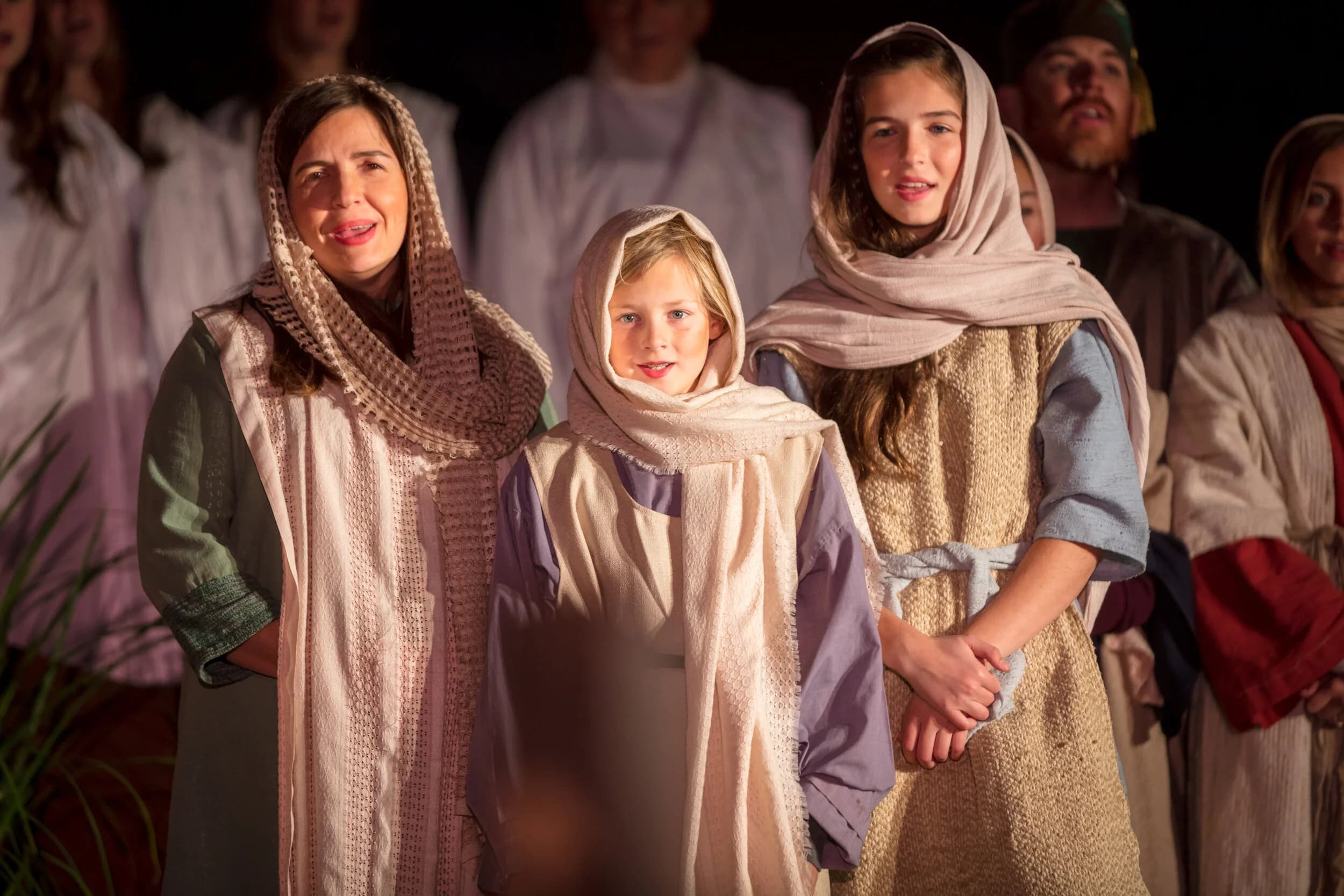 Three girls in headscarves and robes stand together smiling before a crowd of costumed cast members at night.
