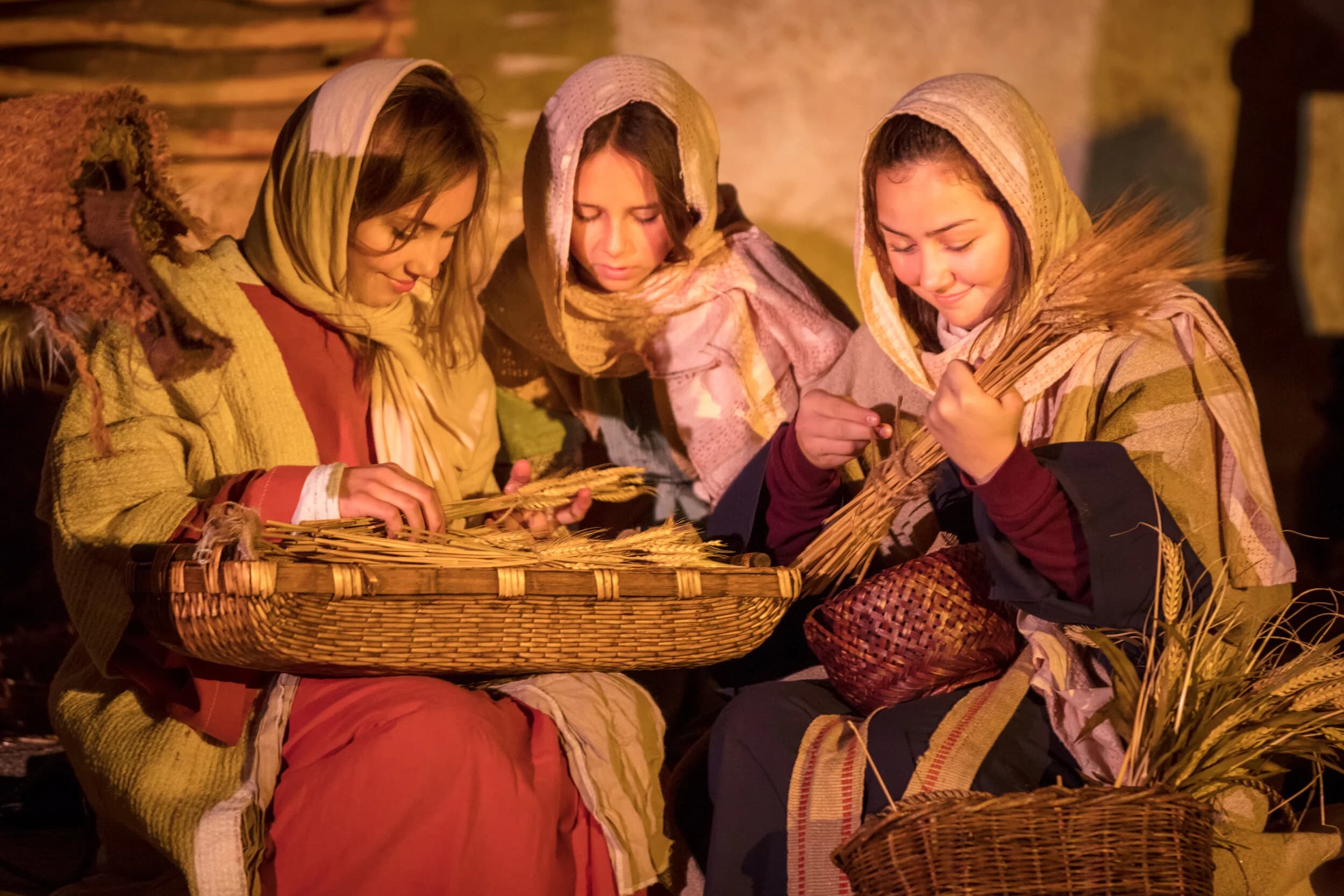 Three young women in headscarves sit together tying bundles of wheat into a woven basket by firelight.