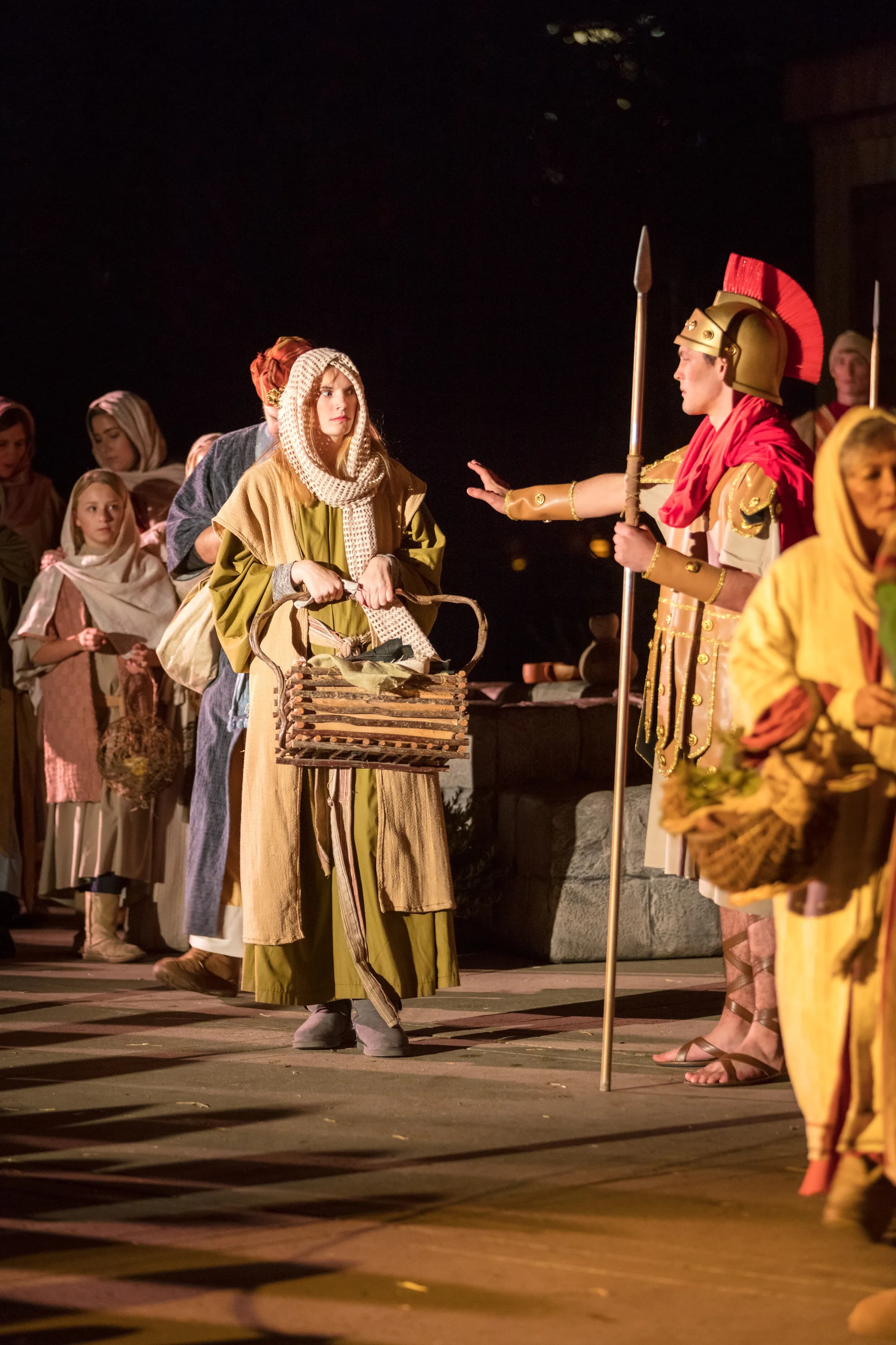 A Roman soldier in a plumed helmet raises his hand toward a young woman carrying a woven basket at a market stall.
