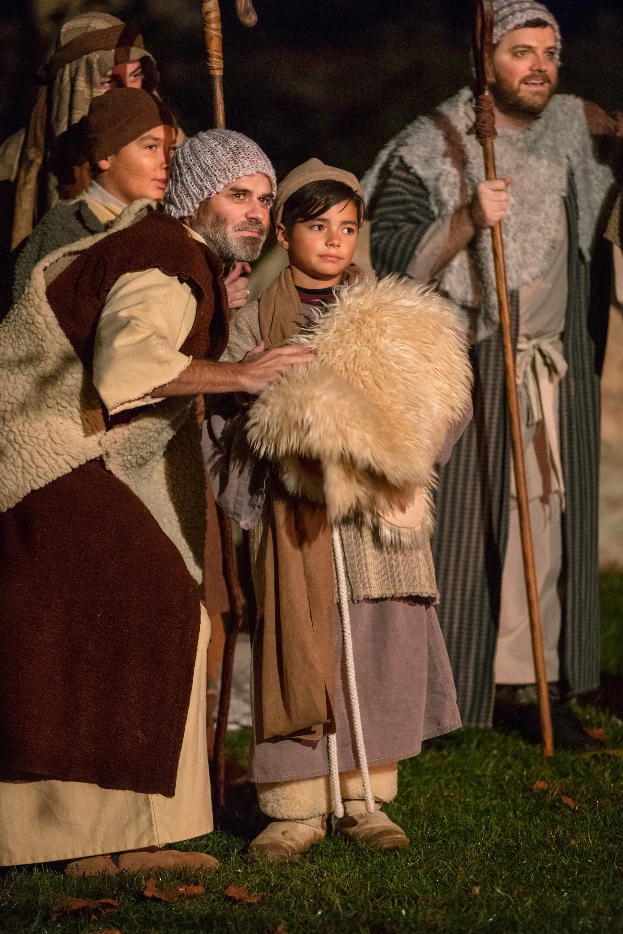 A father and two sons in shepherd robes and knit caps stand together outdoors at night, one boy holding a woolly lamb pelt.