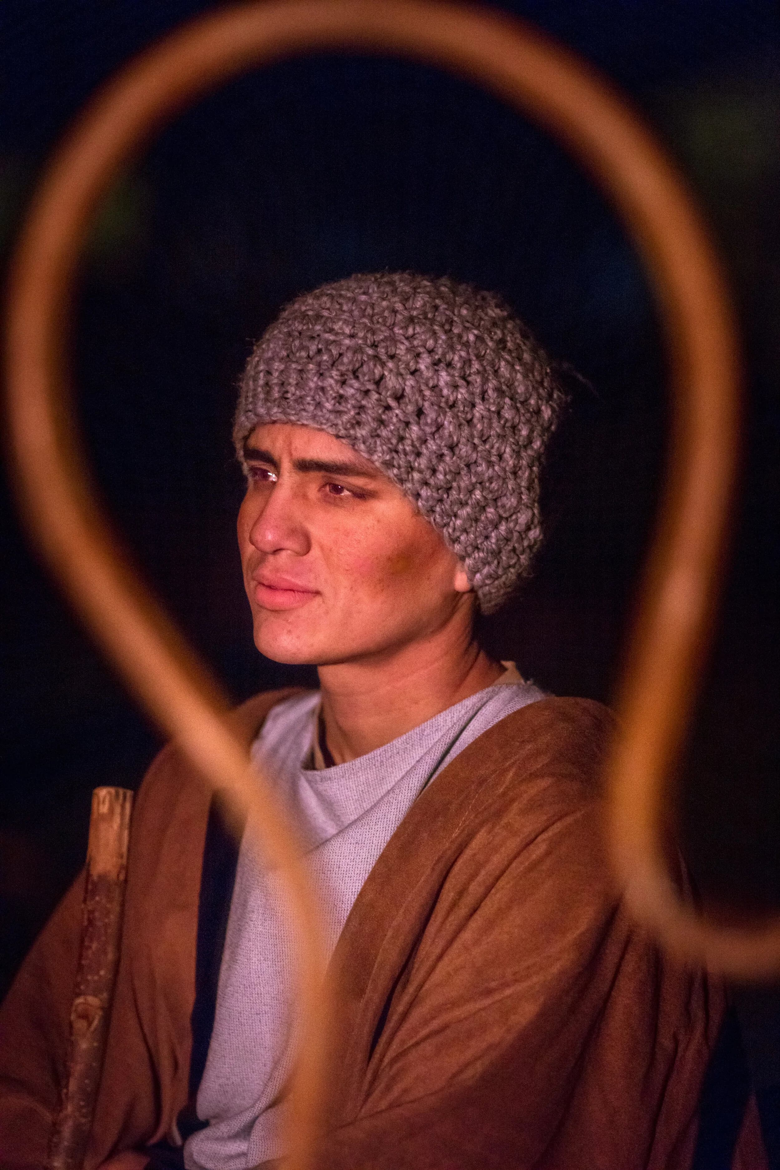 A young man in a knit cap and brown robe looks off to the side, framed by a curved wooden shepherd's crook.