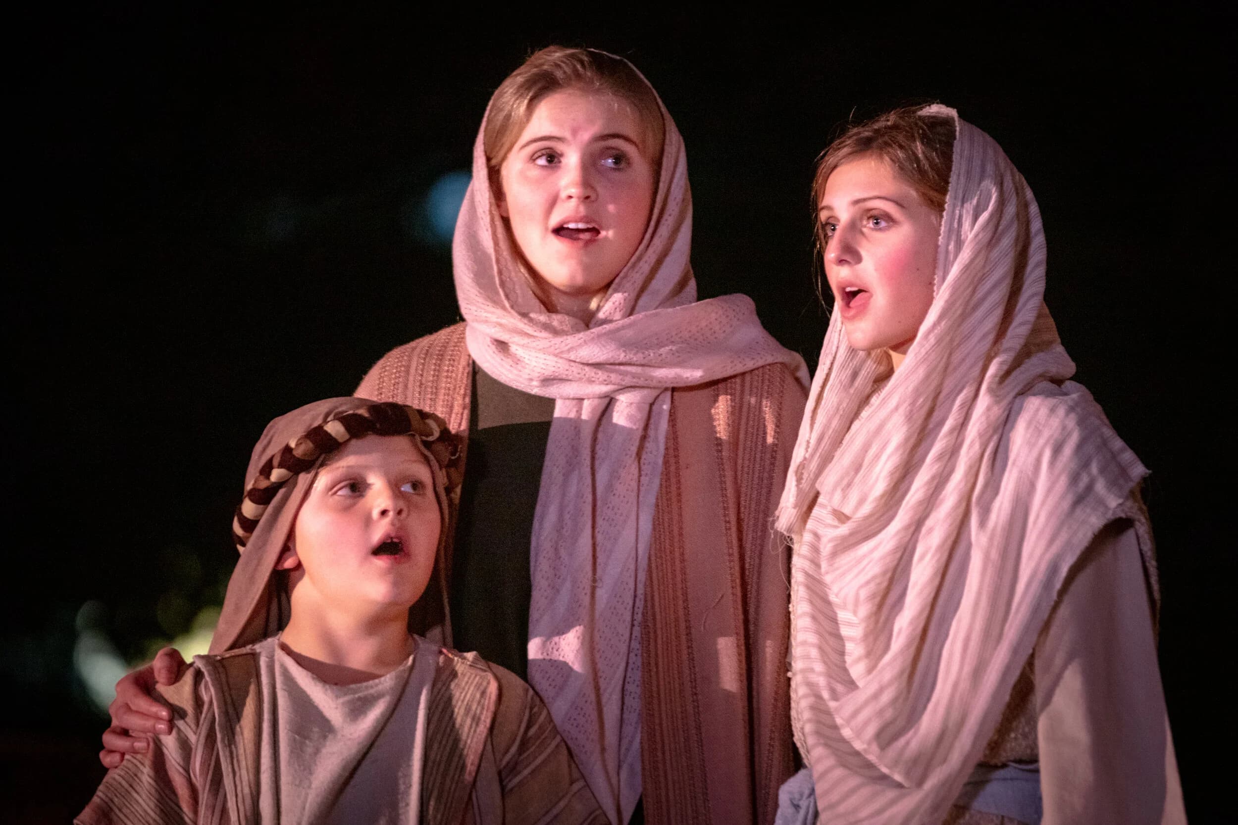 A boy and two teenage girls in headscarves sing together at night, the boy's arm resting on one girl's shoulder.