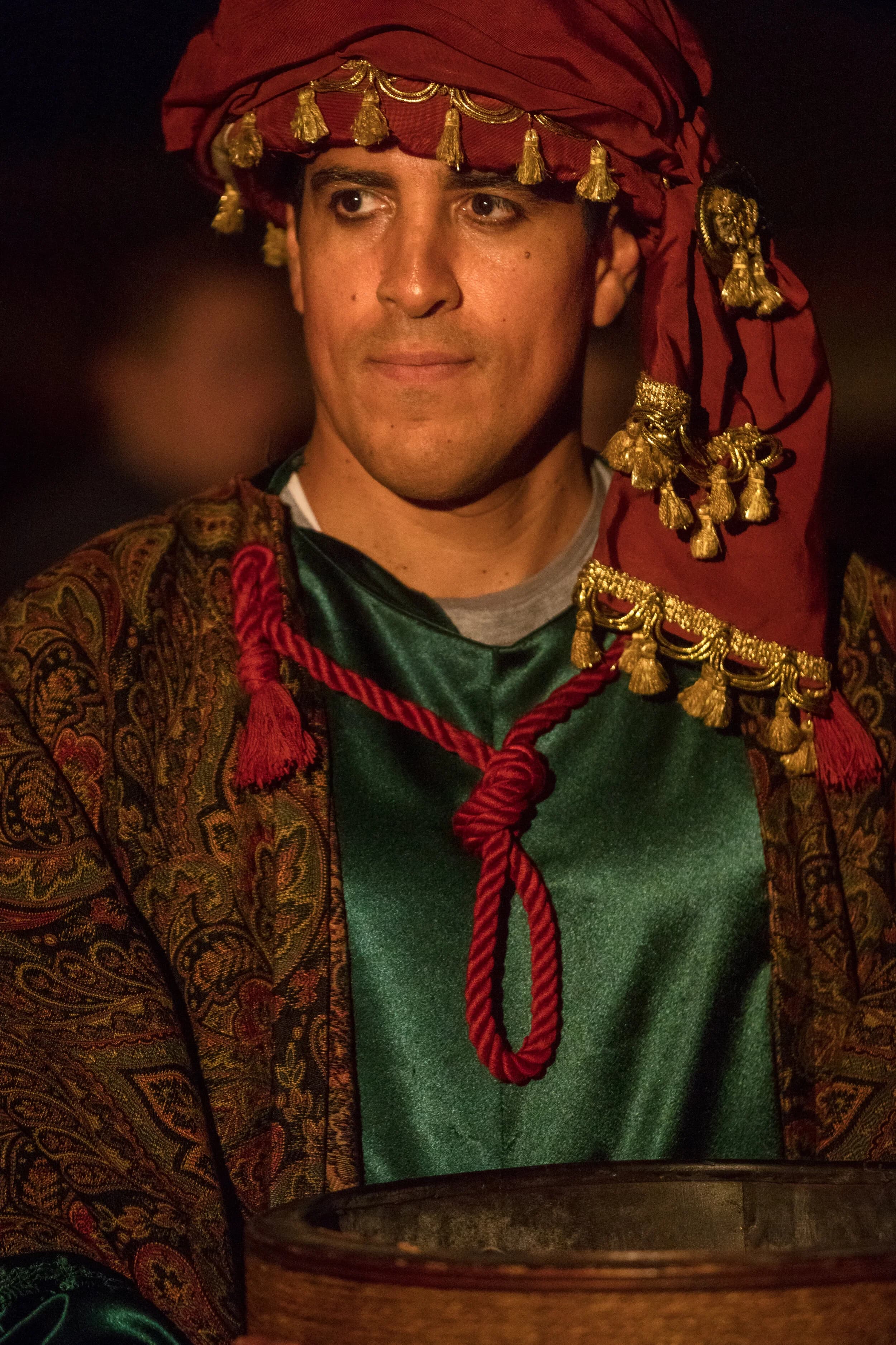 A man wears an ornate red turban trimmed with gold tassels over a green and patterned robe.