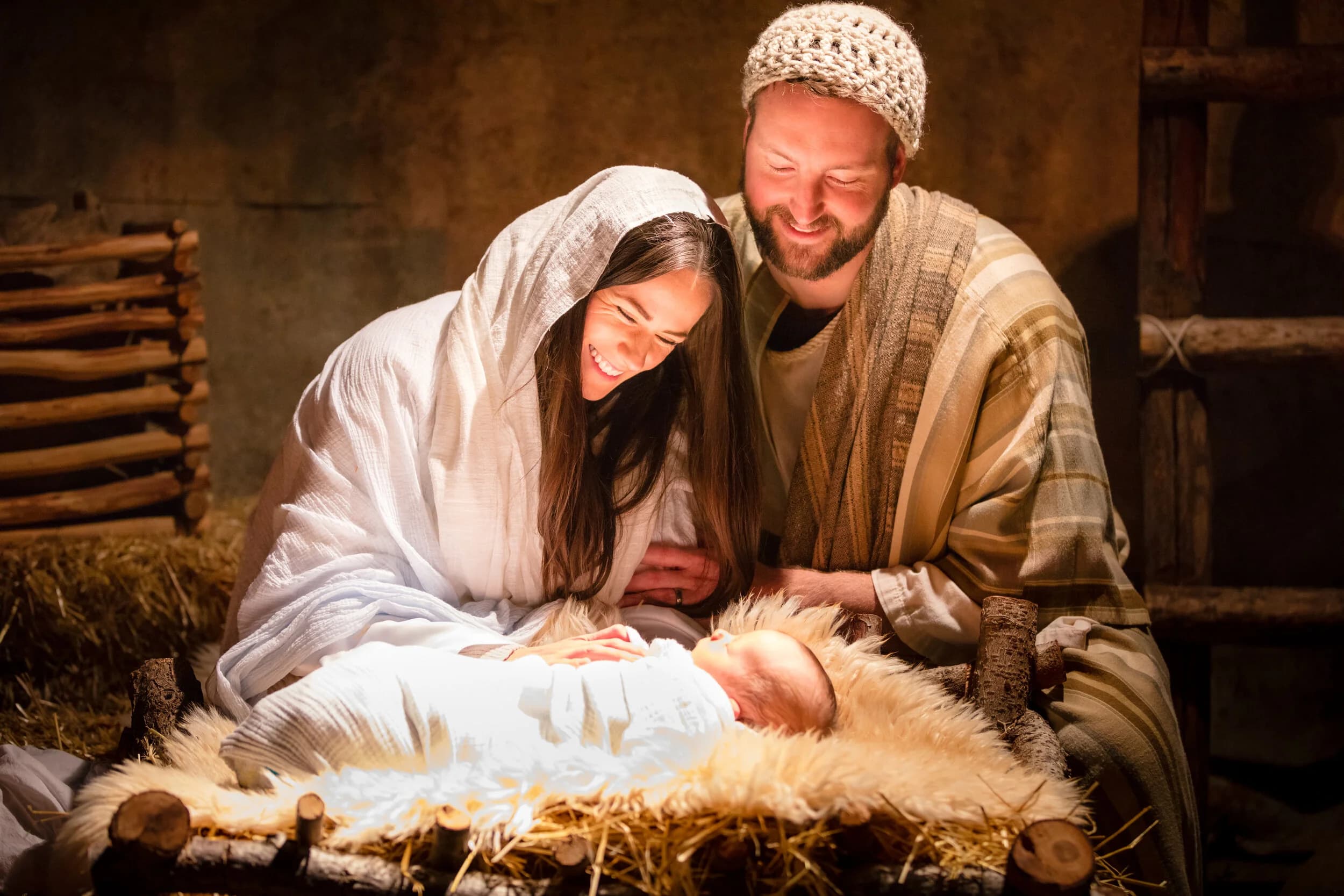 Mary and Joseph kneel together smiling over their baby lying in a fur lined manger at night.