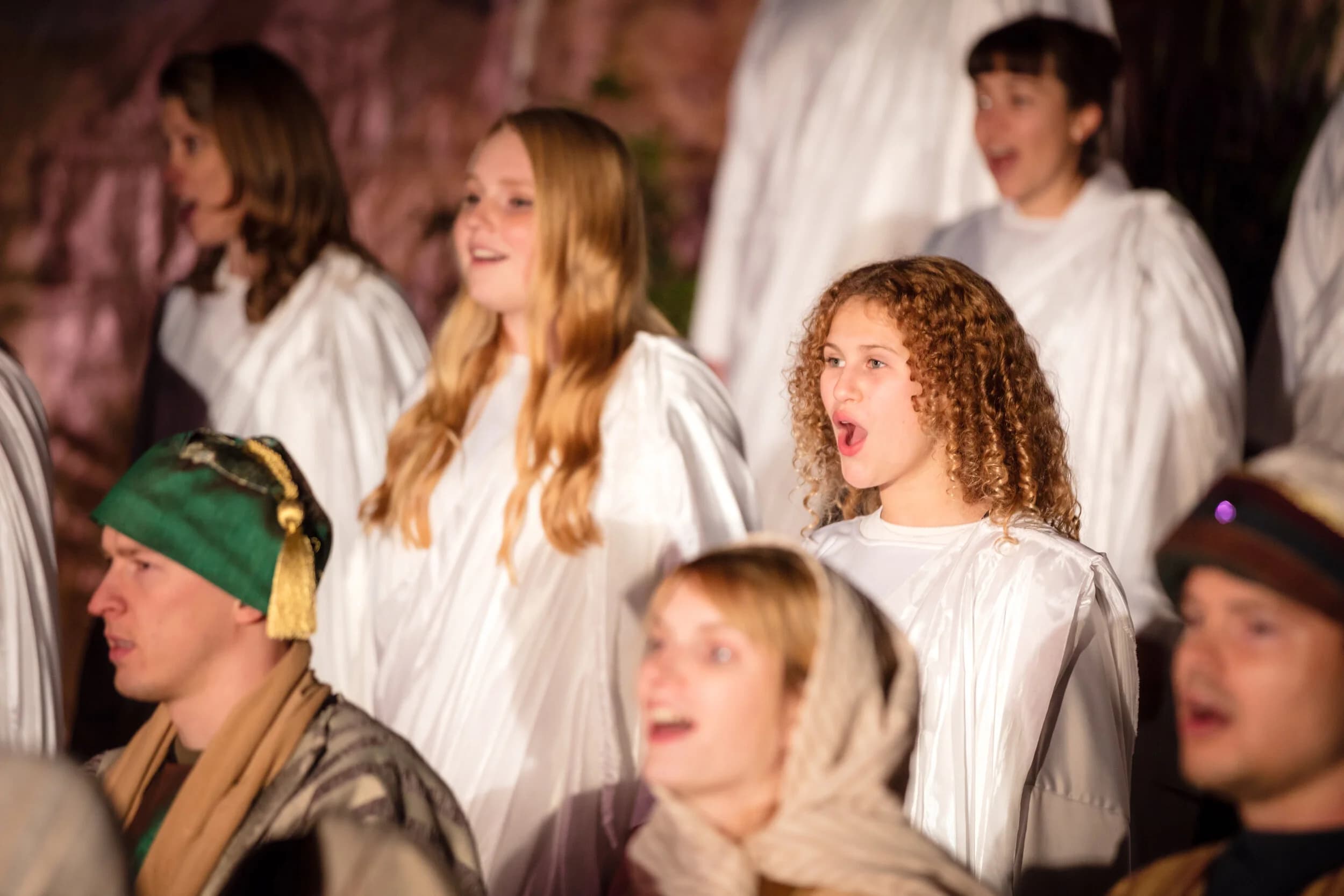 Young women in white choir robes sing together, one with curly hair and her mouth open mid note.