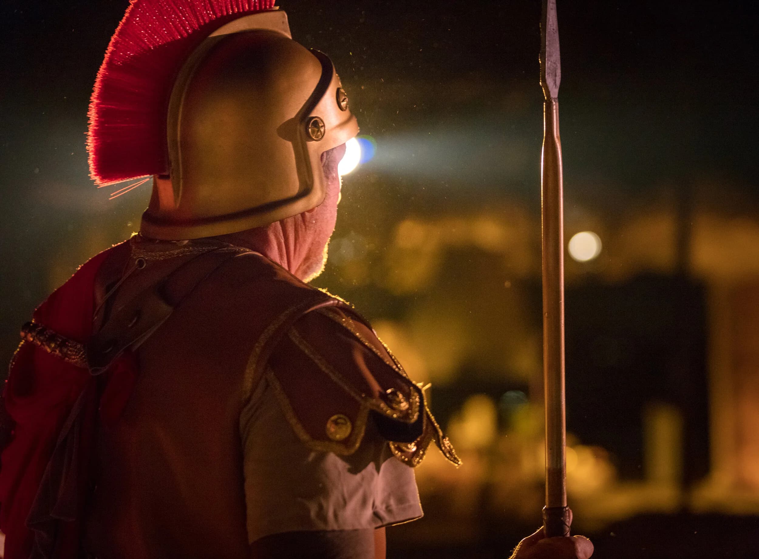 A Roman soldier seen from behind wears a red plumed helmet and cape lit against the night sky, holding a spear.
