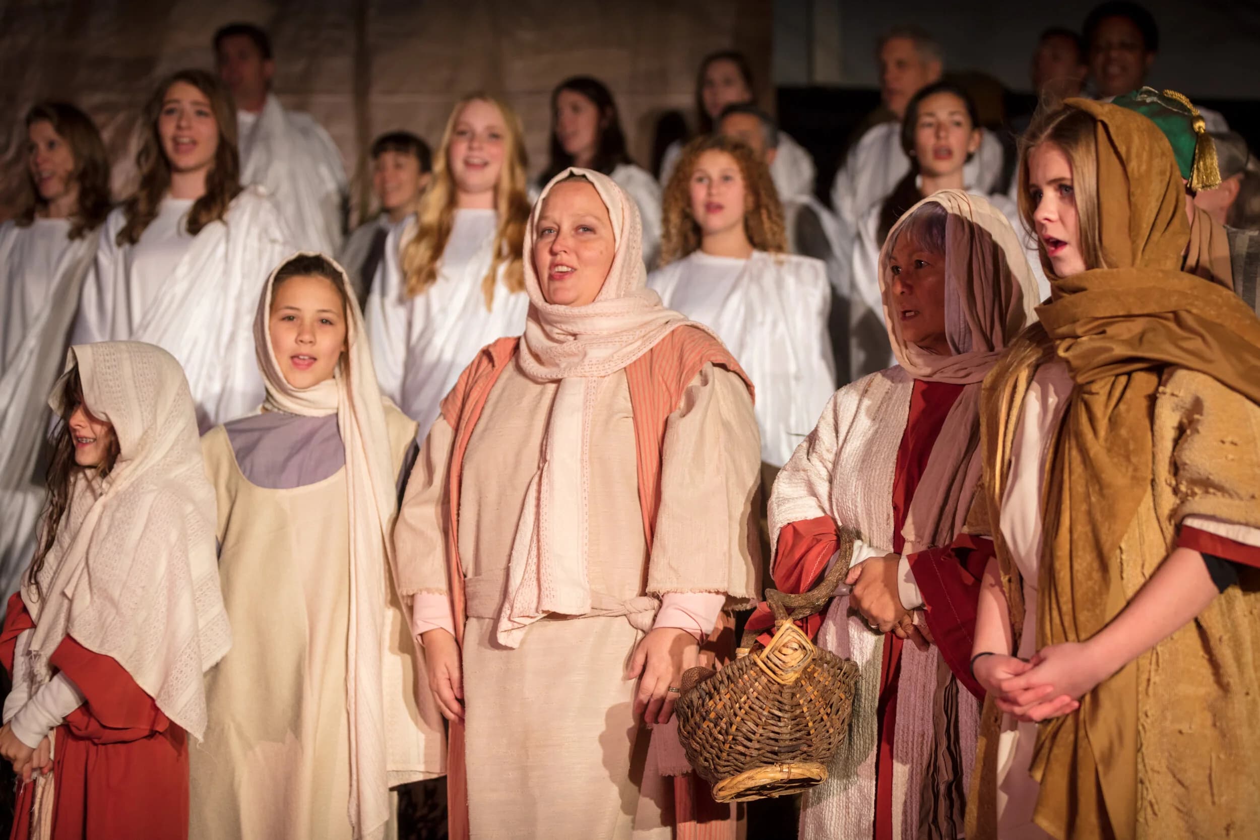 A group of women and girls in headscarves and robes sing together, one holding a woven basket.