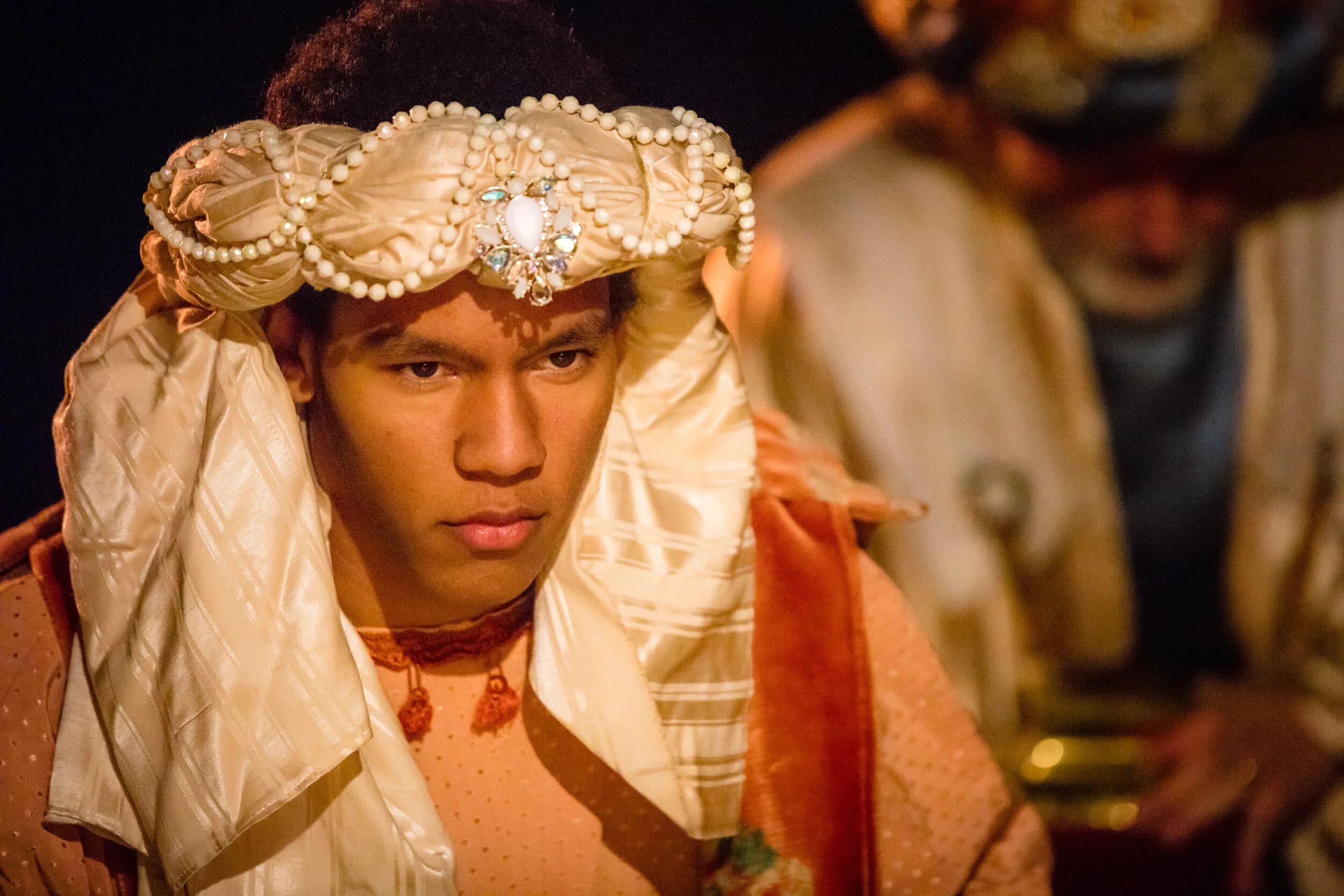 A young man adjusts an ornate pearl and jewel trimmed headpiece over dark curly hair.