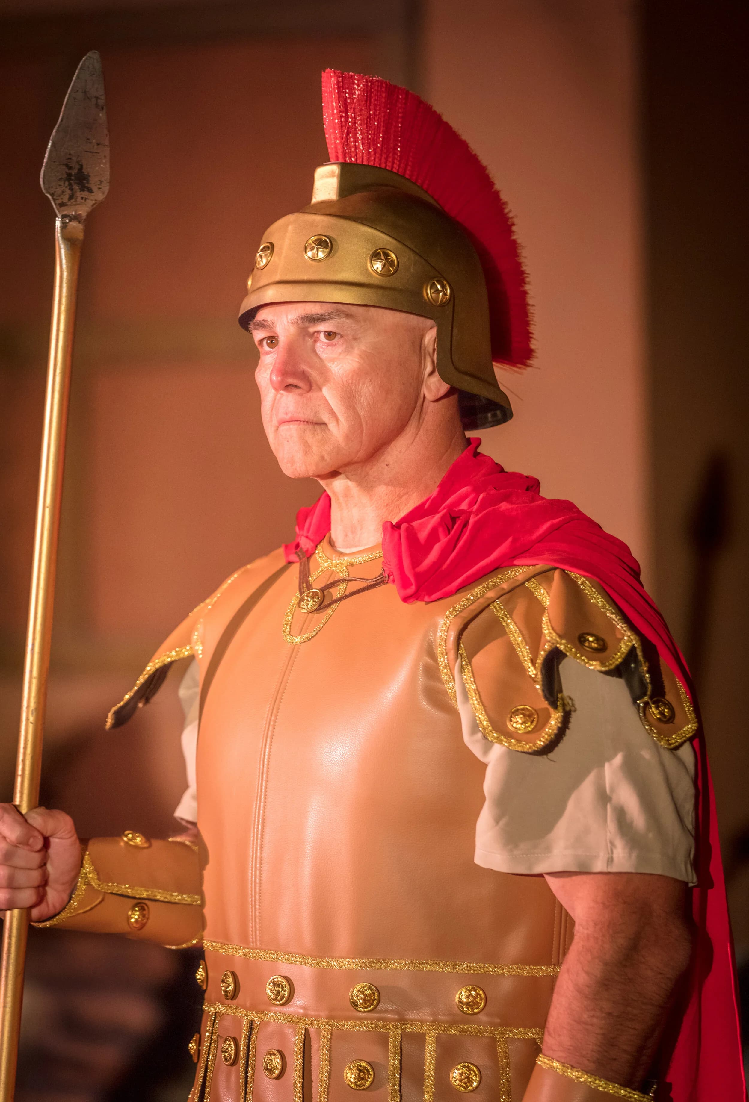 A Roman centurion in a plumed helmet and red cloak stands holding a spear, with gold armor details on his chest.