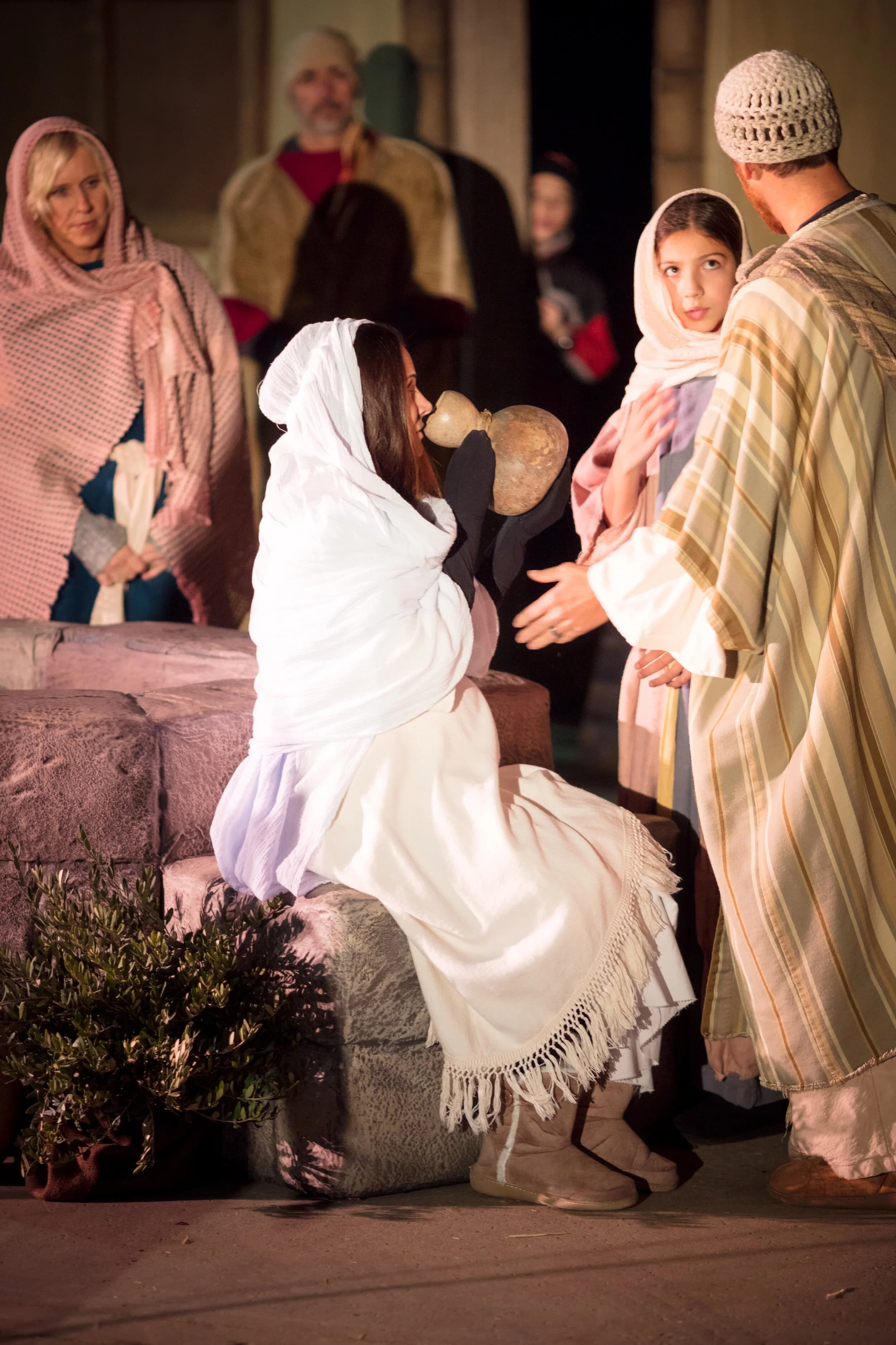 A woman in a white headscarf kneels at a stone well pouring water from a clay jug as others look on.