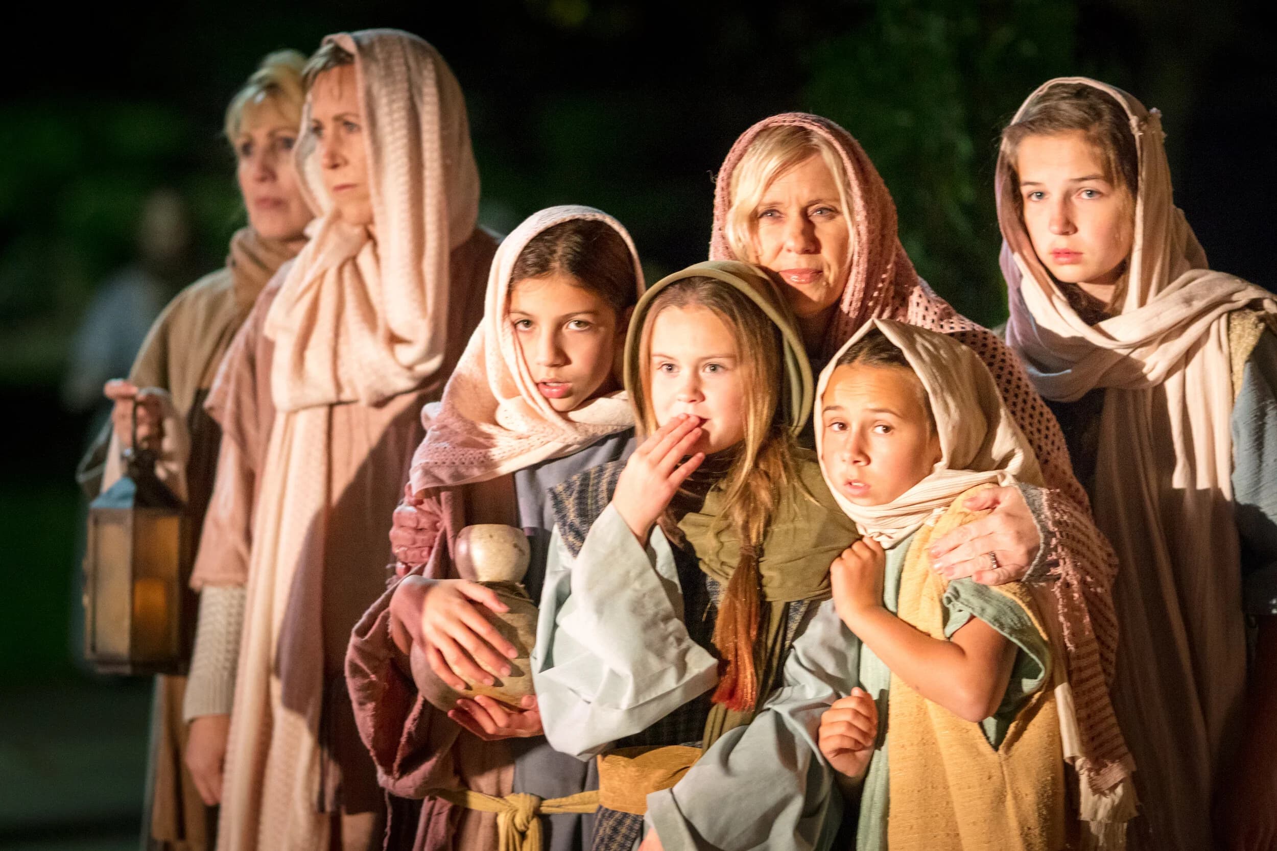 A group of women and girls in headscarves huddle together at night, one holding a small clay jug and a lantern glowing behind them.