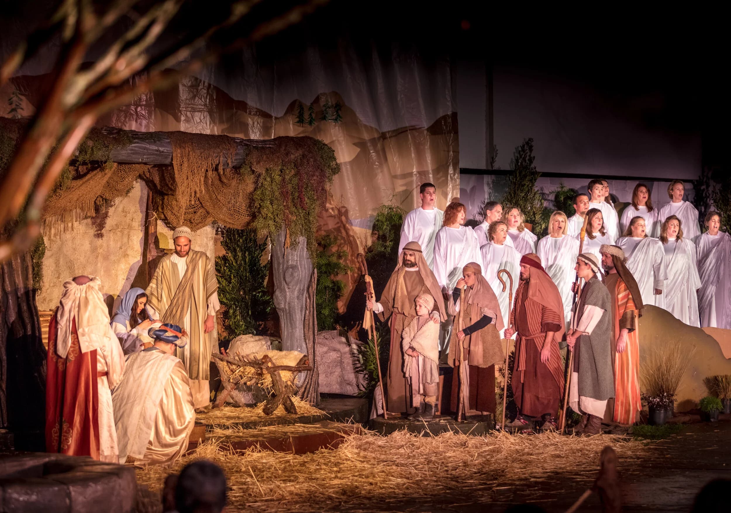 Wise men kneel beside Mary and Joseph at the manger while a choir in white robes and shepherds sing behind them on the stable set.