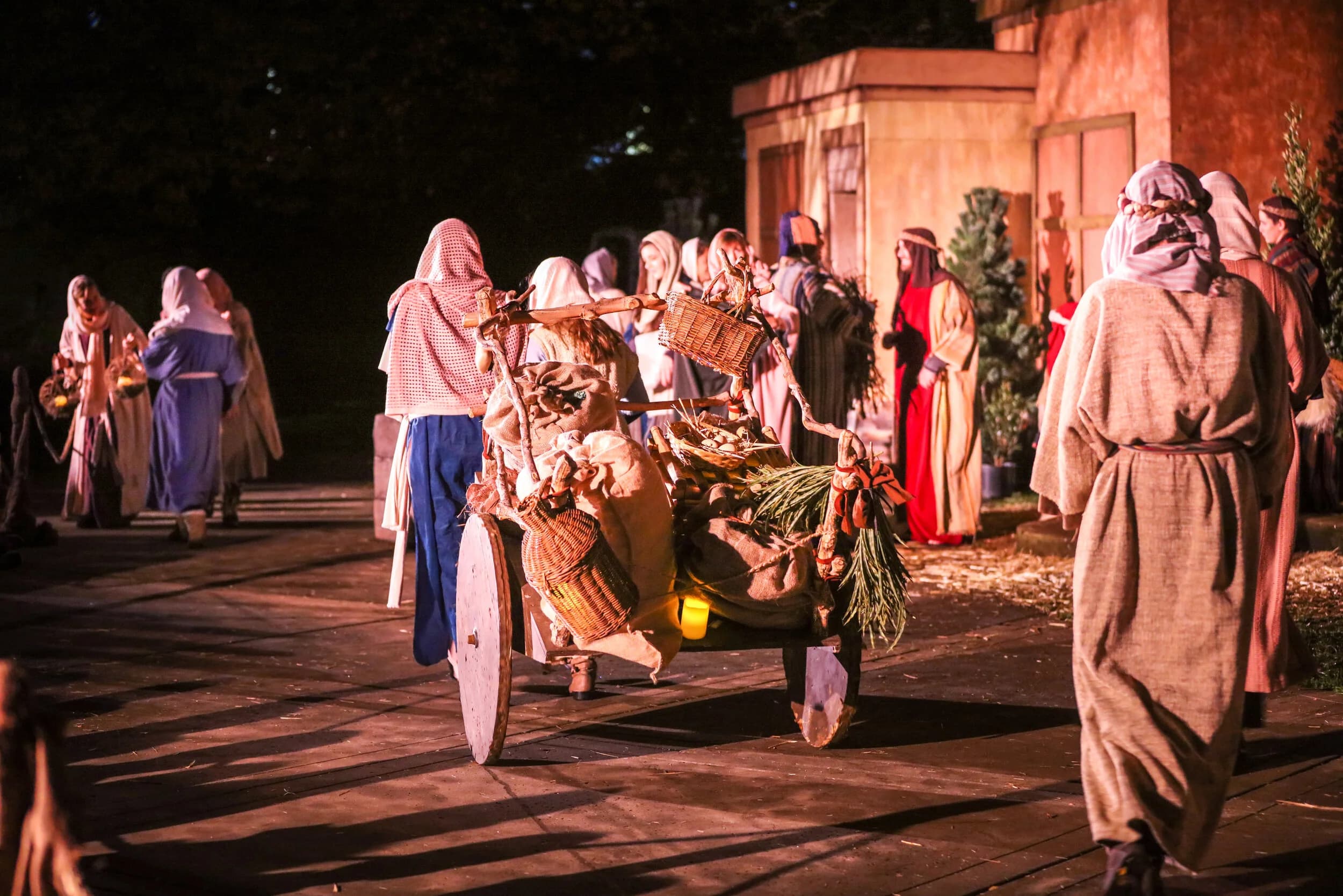 A crowd in robes and headscarves gathers around a small wooden handcart loaded with baskets and bundles outside a lit building at night.