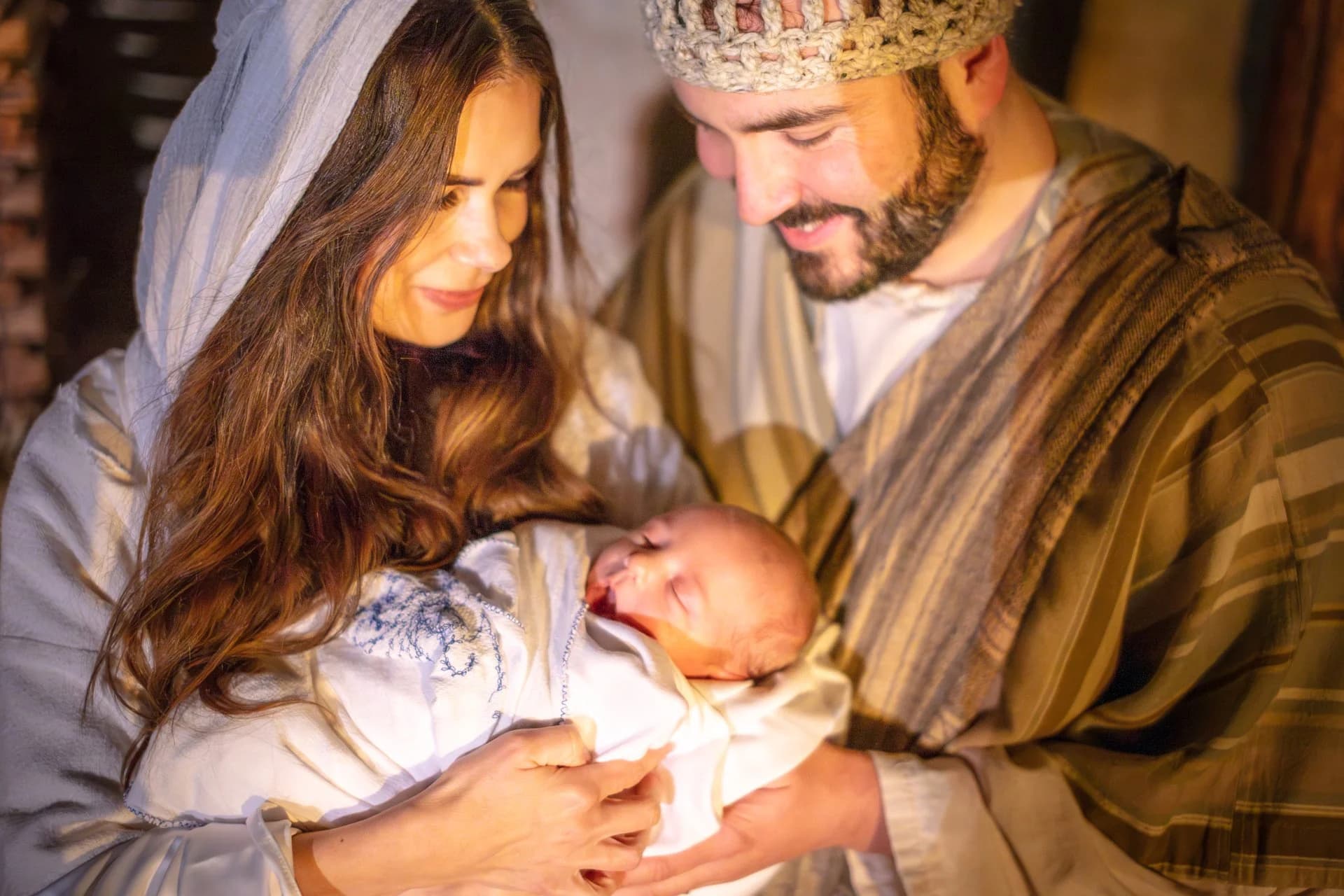 Mary and Joseph lean close over their newborn wrapped in white cloth, lit by warm lantern light.