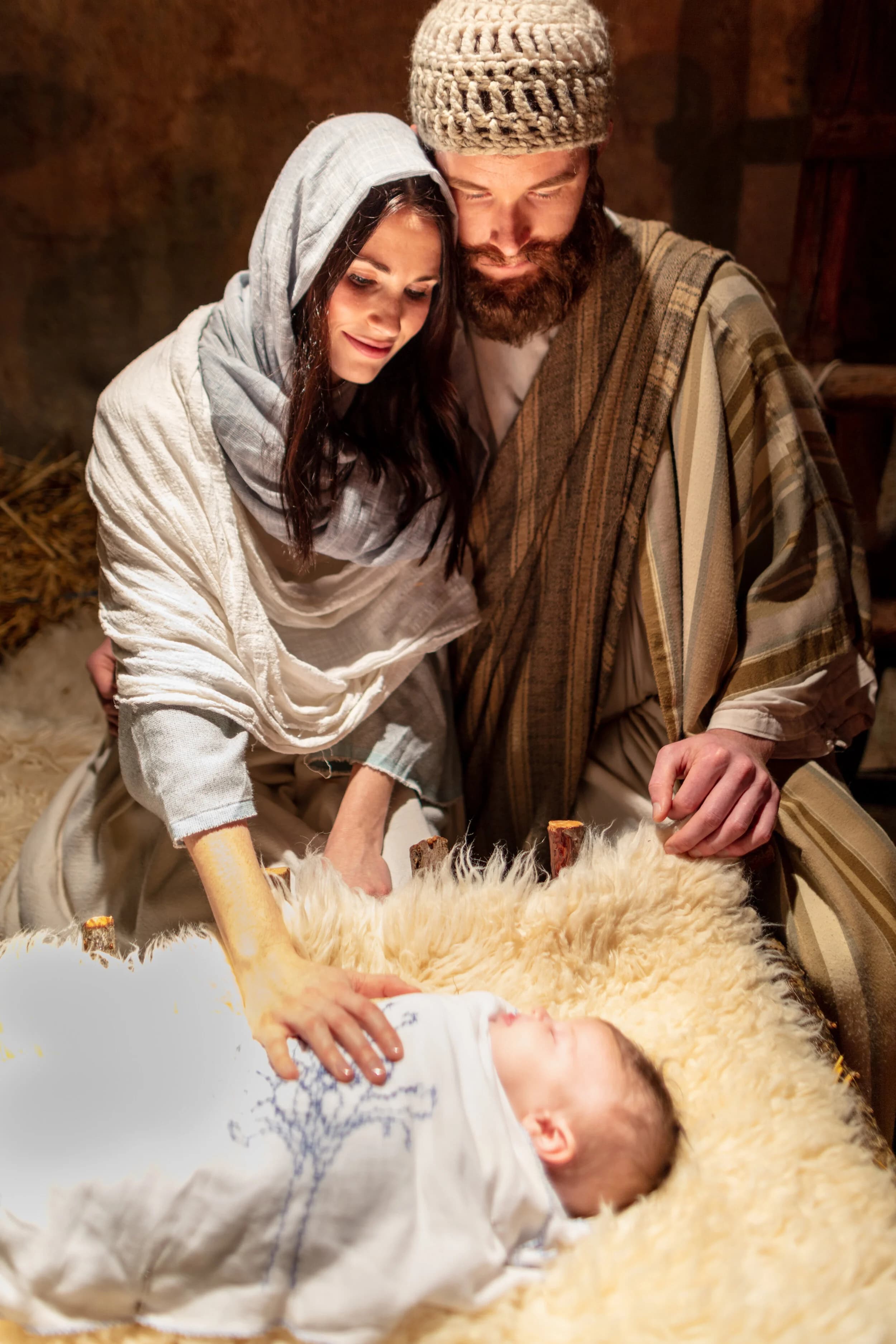 Mary and Joseph kneel together over their newborn laid in a straw filled wooden manger as wise men look on.