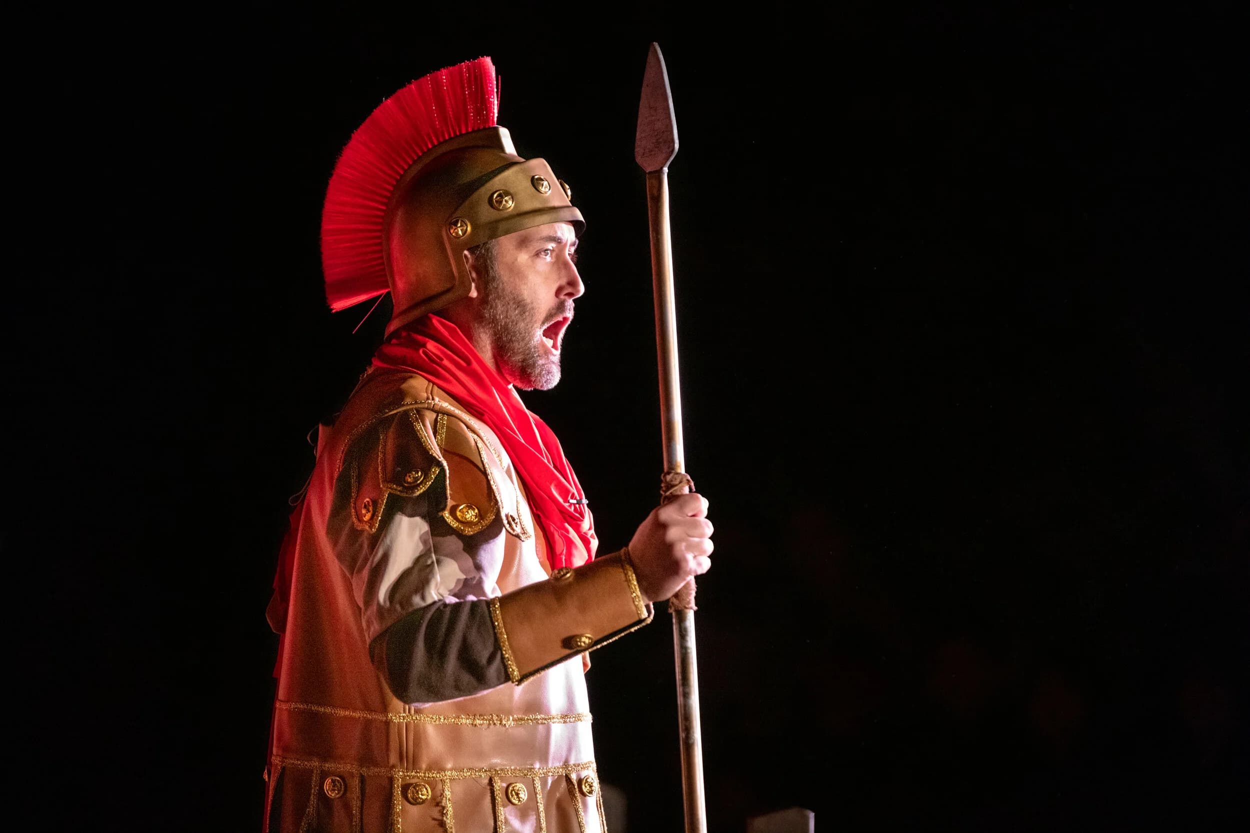 A Roman soldier in profile wears a bronze helmet with a red plume, calling out while holding a spear against a dark background.