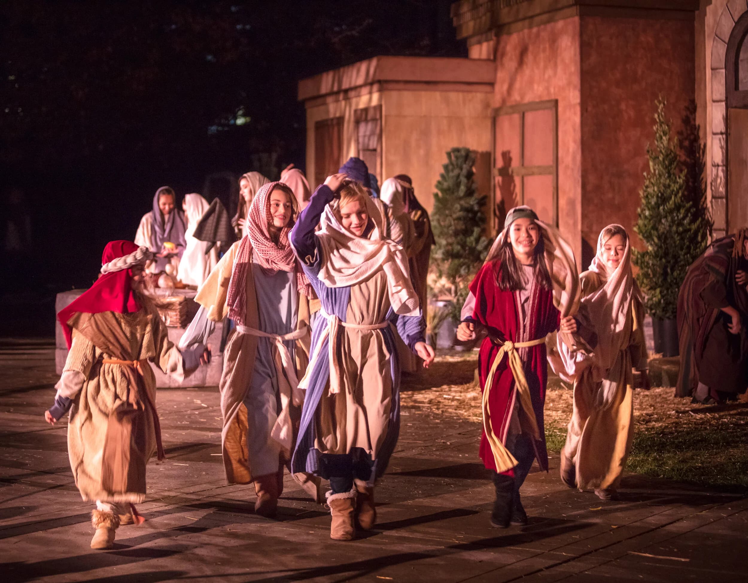 A group of young costumed villagers in robes and headscarves walk arm in arm past market stalls and stone building facades on the torchlit Bethlehem set at night.