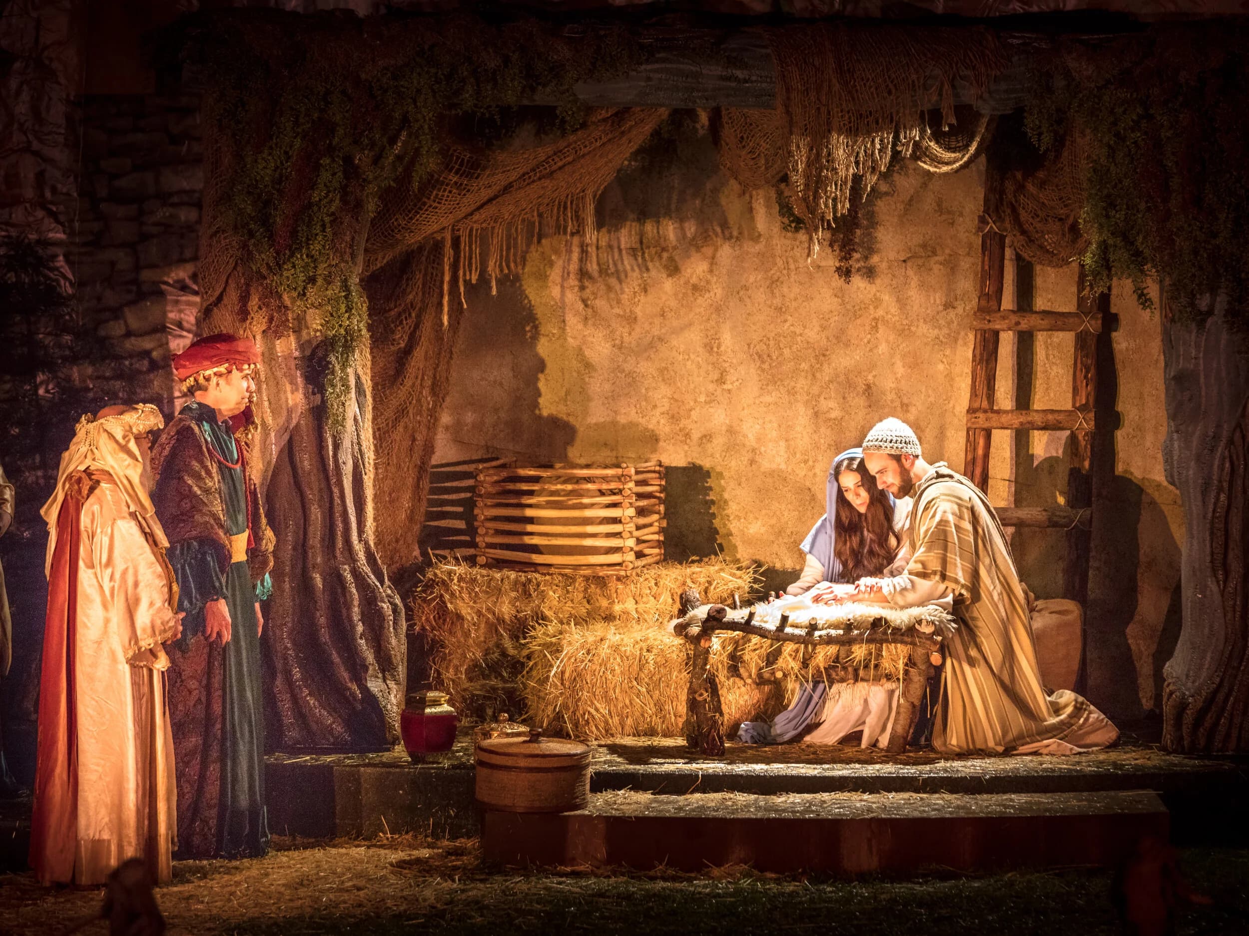 Mary and Joseph kneel beside a wooden manger cradle in a straw-strewn stable while two wise men in richly colored robes look on by warm lantern light.